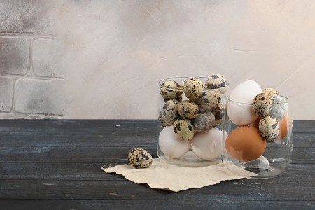 Quail eggs and white and yellow eggs of chicken in a glass container on wooden background.
