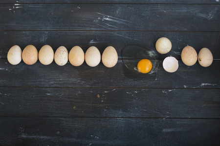 Nine turkey eggs and one broken egg are lying in a row on rough dyed wooden background. Horizontal.