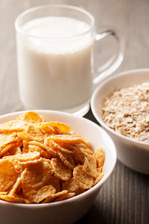 Cornflakes and oatmeal in white bowls with glass of milk on gray wooden table, vertical