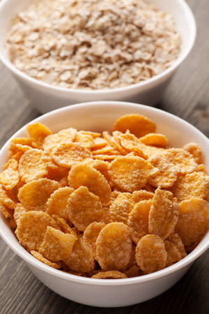 Cornflakes and oatmeal in white bowls on wooden table, vertical