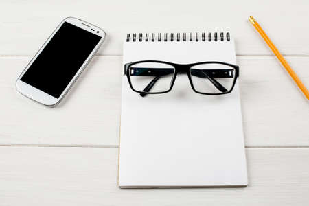 Glasses, notepad, pencil and mobile device on a white wooden table.
