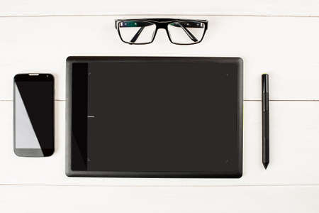 Graphic tablet with eyeglasses and smartphone on a white wooden table.