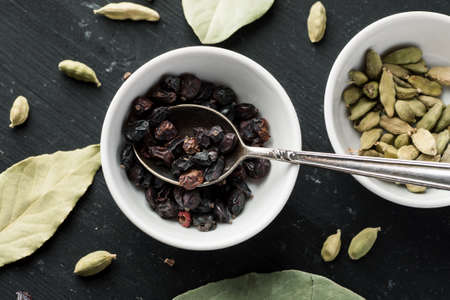 Dry barberry berries in white ceramic bowl with metal spoon in it on a black wooden table with other...