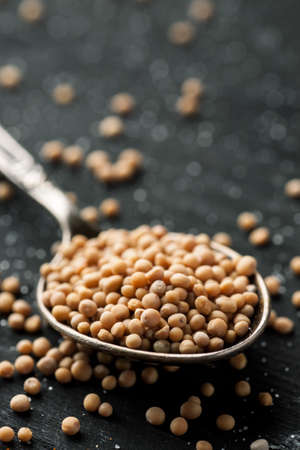 White mustard seeds in metal spoon on a black wooden table, selective focus, vertical, copy space.の写真素材