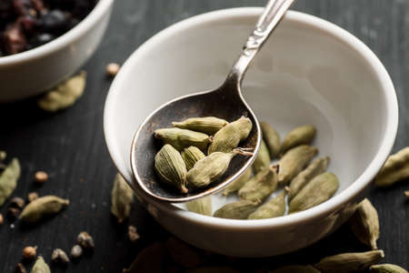 Cardamon seeds in metal spoon above ceramic bowl on a black wooden table.