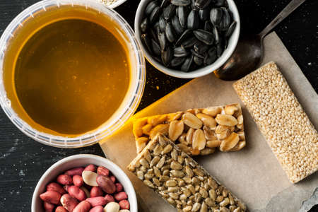 Honey jar, peanuts, sunflower seeds in white ceramic bowls and some brittle on a wooden table, top...