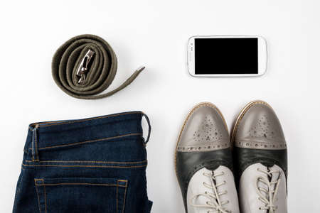 Belt, blue jeans, white smartphone and gray shoes on a white background top view, flat layの写真素材