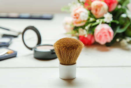 Big cosmetic brush among makeup items on a white wooden table with roses bouquet on the background
