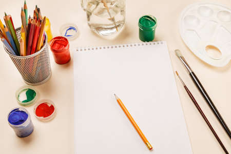 Blank sheet of paper, pencils, paint jars on a table, top viewの写真素材