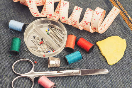 Set of tools and accessories for sewing and needlework with threads in spools, needles, measuring tape and other items on piece of jeans fabricの写真素材