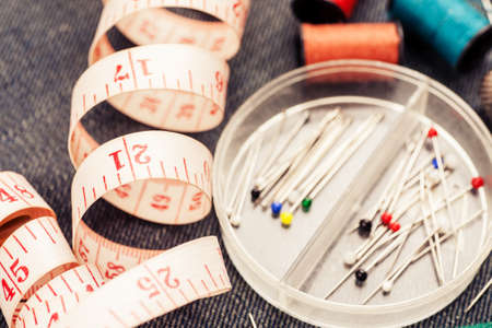 Set of tools and accessories for sewing and needlework with threads in spools, needles, measuring tape and other items on piece of jeans fabricの写真素材
