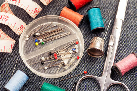 Set of tools and accessories for sewing and needlework with threads in spools, needles, measuring tape and other items on piece of jeans fabricの写真素材