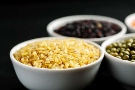 Set of different cereals and legumes in a white bowls on a black tableの写真素材