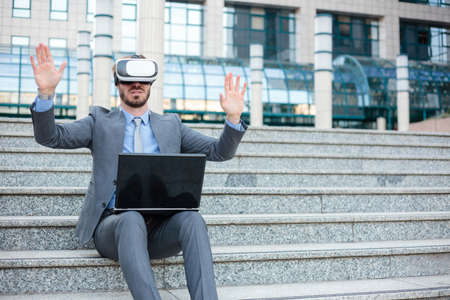 Handsome young businessman using VR goggles and making hand gestures, working on a laptop in front of an office building. Working with modern technologies conceptの写真素材