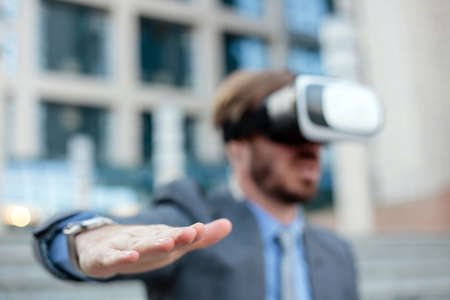 Close up of a young businessman using VR goggles in front of an office building, making hand gestures. Selective focus on his hand in foreground. Working with modern technologies conceptの写真素材