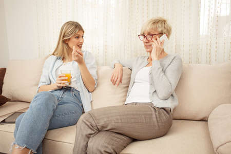 Young woman is curious and anticipating good news from her mother who is talking on the phone. Happy family moments at homeの写真素材