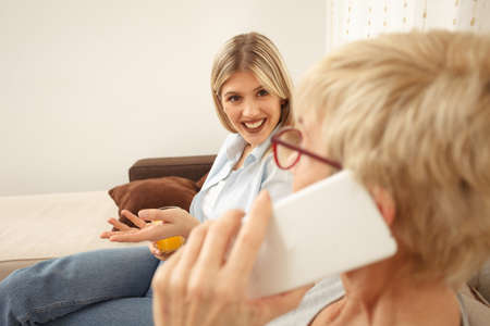 Young woman is curious and anticipating good news from her mother who is talking on the phone. Focus on daughter in background. Happy family moments at homeの写真素材