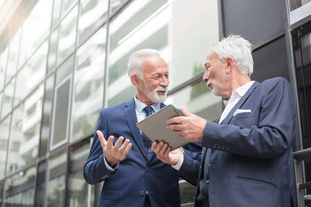 Two smiling senior gray haired businessman working a tablet and discussing. Standing in front of an office building. Low angle viewの写真素材