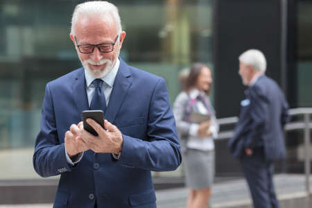 Successful happy senior gray haired businessman using smart phone, browsing internet or messaging. Two people talking in the background, blurredの写真素材
