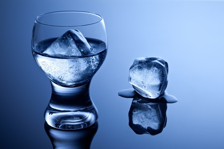 An elegant shot glass with vodka and ice along with a melting ice cube on reflective surface.の写真素材