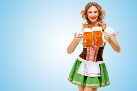 Young sexy Oktoberfest woman wearing a traditional Bavarian dress dirndl serving two beer mugs with happy smile on blue background の写真素材