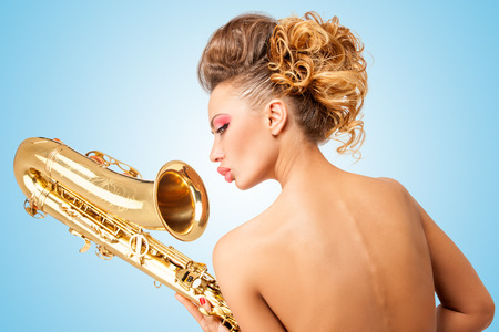 Retro portrait of a nude pin-up girl with vintage winged eyes, turning her back on and holding a saxophone upside down on blue background.の写真素材