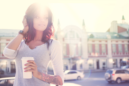 Beautiful young smiling woman with tattoo, holding a disposable takeaway cup and looking aside against urban city background.の写真素材