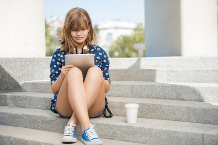 Beautiful young woman with vintage music headphones around her neck and a take away coffee cup, surfing internet on tablet pc and sitting on stairs against urban city background.の写真素材