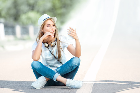 Happy young woman with vintage music headphones around her neck, surfing internet on a smartphone and sitting on a separating strip against road background.の写真素材
