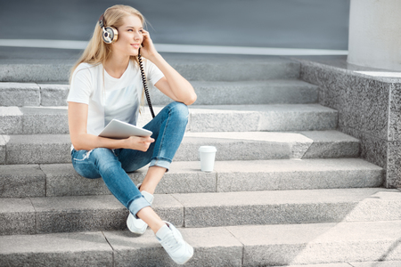 Happy young woman with vintage music headphones and a take away coffee cup, surfing internet on tablet pc, listening to the music and sitting on stairs against urban city background.の写真素材