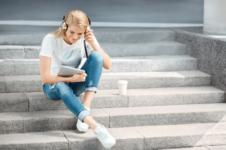Happy young woman with vintage music headphones and a take away coffee cup, surfing internet on tablet pc, listening to the music and sitting on stairs against urban city background.の写真素材