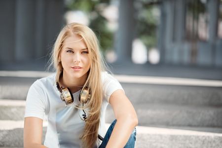 Beautiful young woman with vintage music headphones around her neck, sitting against urban city background and looking straight.の写真素材