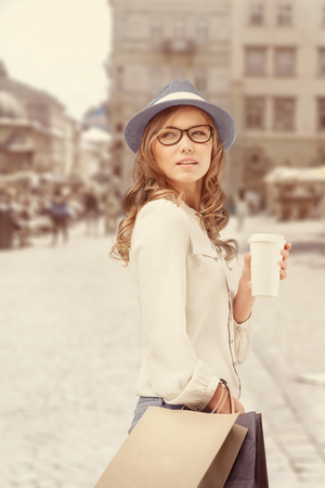 Happy young fashionable woman with bags having a coffee break after shopping and holding take away coffee against urban background.の写真素材