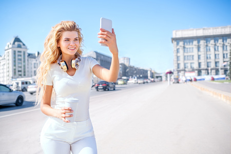 Beautiful young woman with music headphones and a take away coffee cup, taking picture of herself, selfie against urban city background.の写真素材