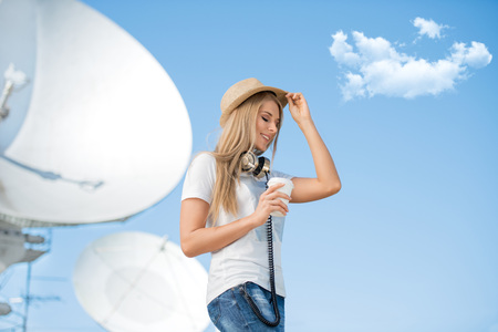 Happy young woman in hat wearing vintage music headphones around her neck, drinking takeaway coffee and posing against background of parabolic satellite dish that receives wireless signals from satellites.の写真素材