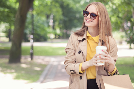Young fashionable woman taking a coffee break after shopping, walking with a coffee-to-go in her hands against green city park background.の写真素材