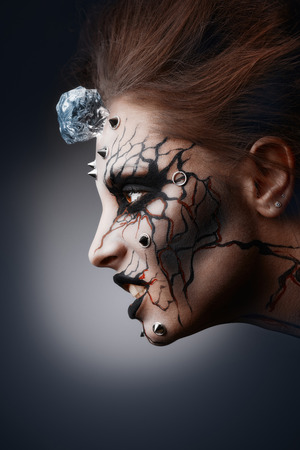 The pierced female face painted as a grinning halloween witch with a cracked paint and an ice cube.の写真素材