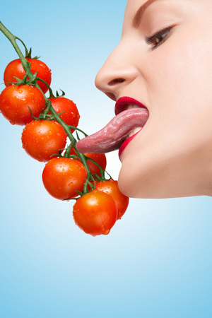 Beautiful girl licking ripe red cherry tomatoes on the vine sexually with her seductive tongue on blue background.の写真素材