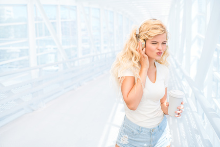 Beautiful young woman with music headphones, standing on the bridge with a take away coffee cup and posing against urban background.の写真素材