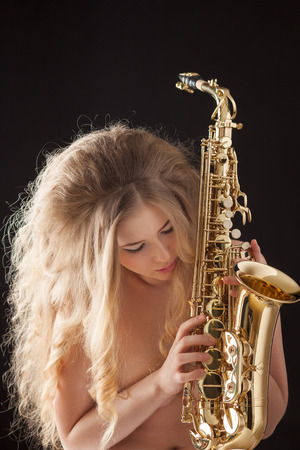 Portrait of a sexy beautiful nude pin-up girl from 60s or 70s, posing with a gold jazz saxophone and pressing keys on black background.の写真素材