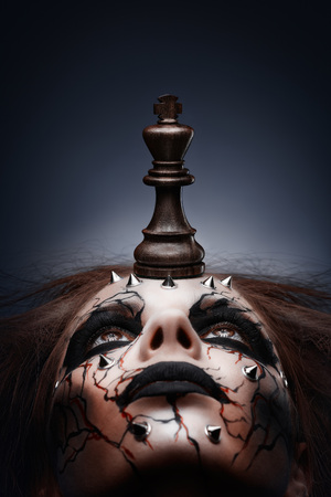 A creative photo of a queen with a painted pierced face and a chess king in her mouth.の写真素材