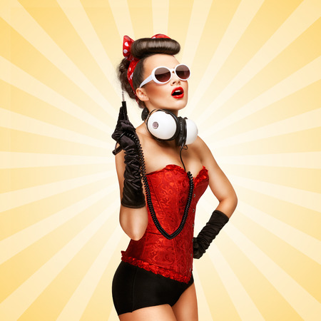 Retro photo of a glamorous pin-up girl with big vintage unplugged music headphones on colorful abstract cartoon style background.の写真素材