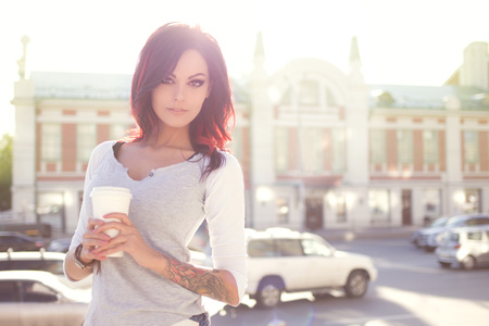 Beautiful young woman in a modern trench coat, holding a disposable takeaway cup and standing against urban city background.の写真素材