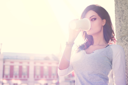 Beautiful young woman against urban concrete wall background drinking coffee from a disposable takeaway cup and looking aside.の写真素材