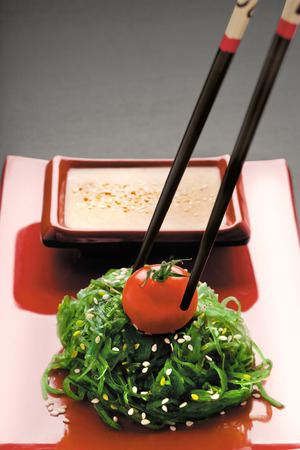 A close-up of chopsticks holding a red-ripe tomato on chuka salad along with gomadare sauce in the background.の写真素材