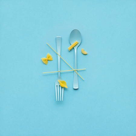 Creative still life photo of fork and spoon with raw pasta on blue background.の写真素材