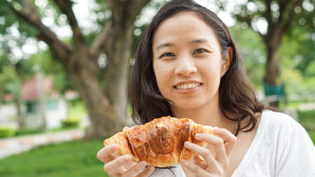 Asian woman holding and eating fresh baked bread bakery in green background parkの写真素材