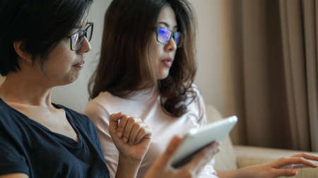 Two Asian friends watching thing on tablet and computer sharing idea at homeの写真素材