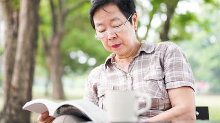 Asian senior woman reading book drinking coffee in sunny parkの写真素材
