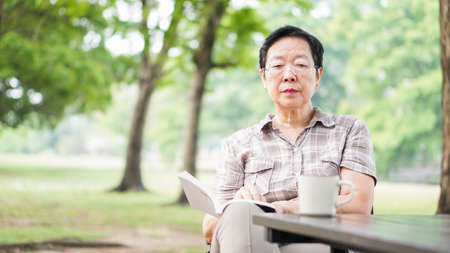 Asian senior woman reading book drinking coffee in sunny parkの写真素材